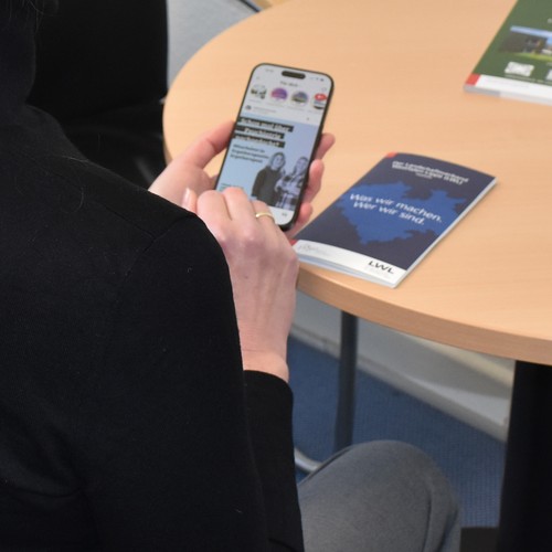 Frau mit schwarzem Pullover sitzt vor einem hellbraunen Tisch und schaut in ihr Handy. Frau mit schwarzem Pullover sitzt vor einem hellbraunen Tisch und schaut in ihr Handy.