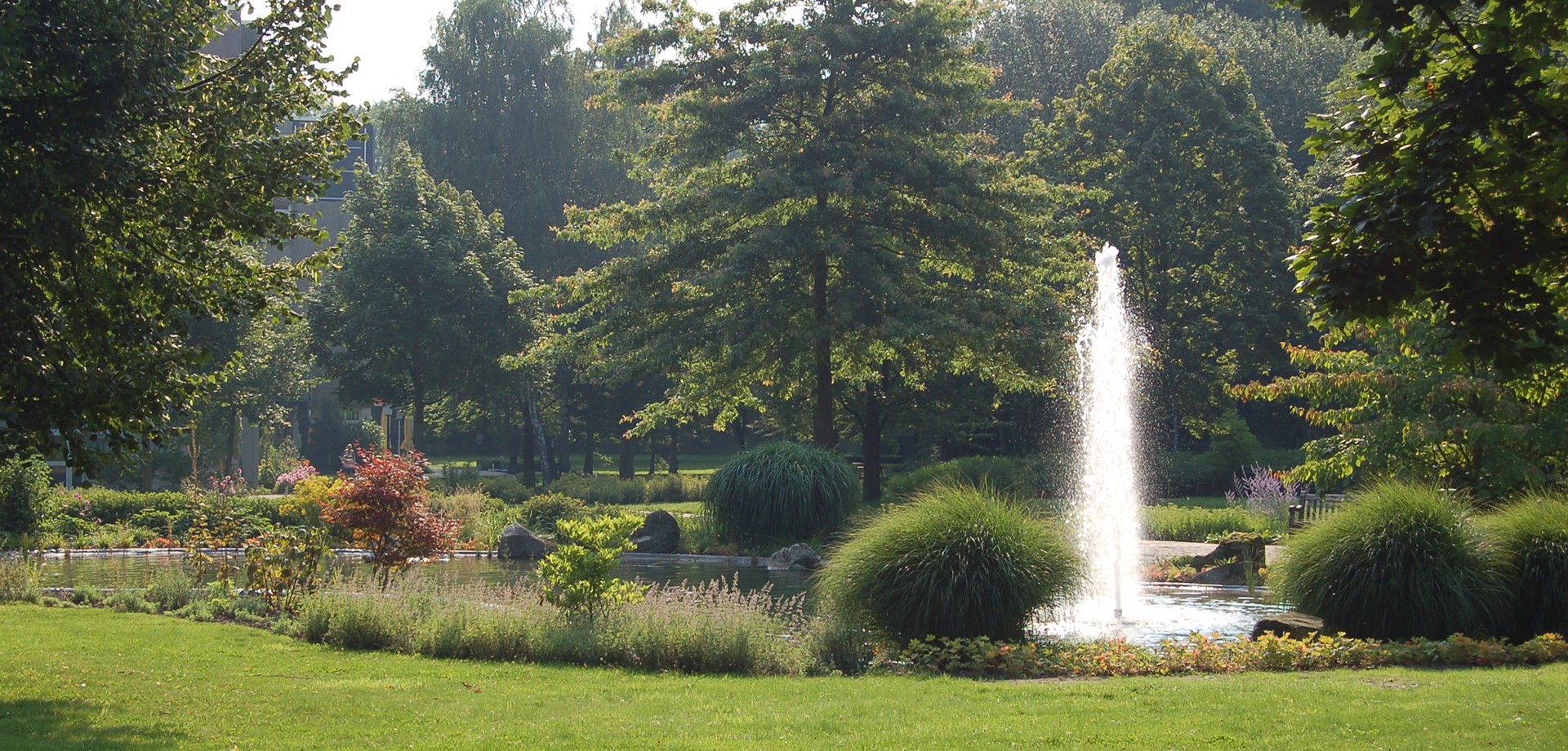 Springbrunnen mit Fontäne inmitten der grünen Parklandschaft der Klinik mit hohen Bäumen im Sommer