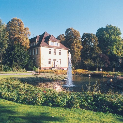 Blick in den grünen Klinikpark, in der Mitte steht ein weißes Haus, davor liegt ein kleiner Teich.