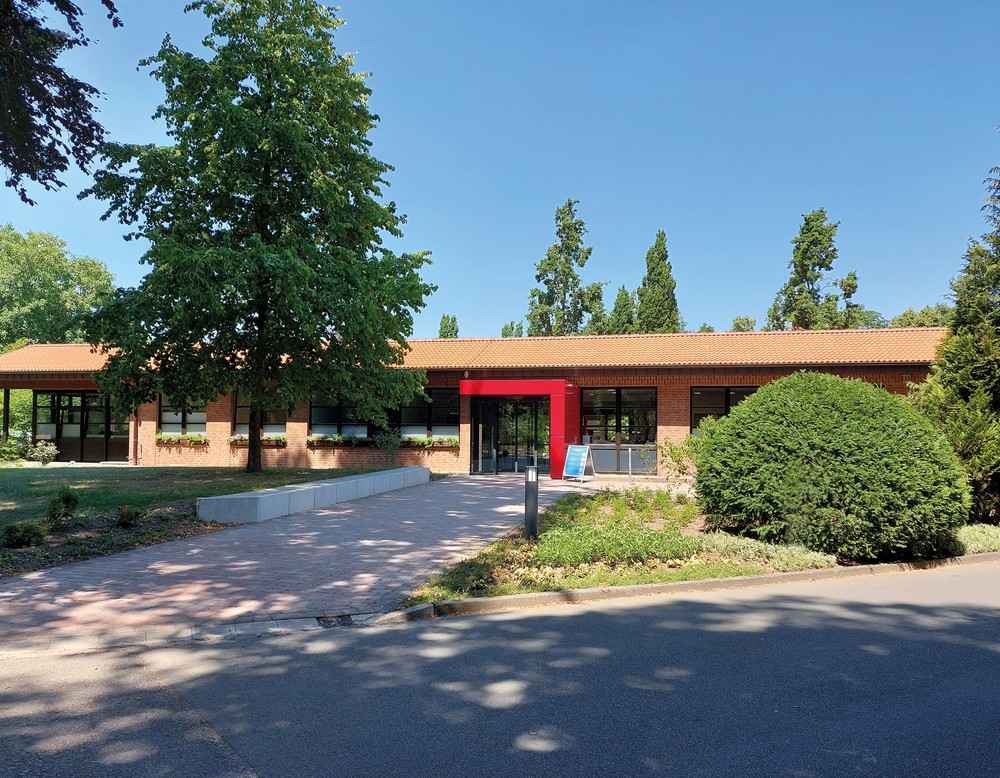 Gebäude der Institutsambulanz mittig im Bild, Haus ohne weitere Stockwerke mit rot eingerahmter Tür, hellbraunes Dach, umrahmt von grünen Bäumen