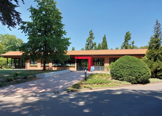 Ambulanzgebäude der Klinik mittig im Bild, Gebäude ohne Stockwerke, mit rotem Stein umrahmter Eingangstür, hellbraunes Dach, umrahmt von grünen Bäumen Ambulanzgebäude der Klinik mittig im Bild, Gebäude ohne Stockwerke, mit rotem Stein umrahmter Eingangstür, hellbraunes Dach, umrahmt von grünen Bäumen