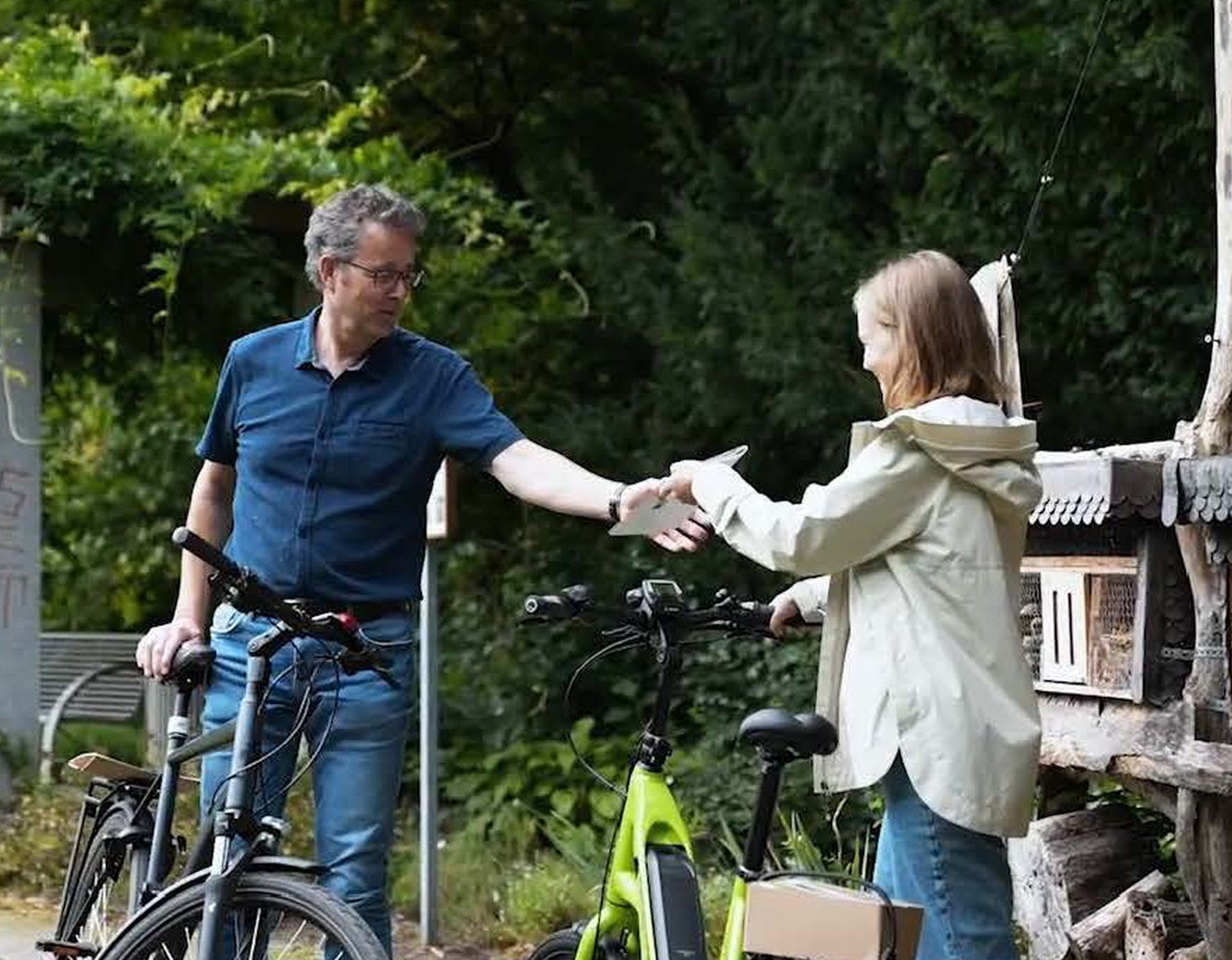 Ein Mann und eine Frau stehen sich mit einem Fahrrad in einem grünen Park gegenüber, die Frau gibt dem Mann eine Broschüre.