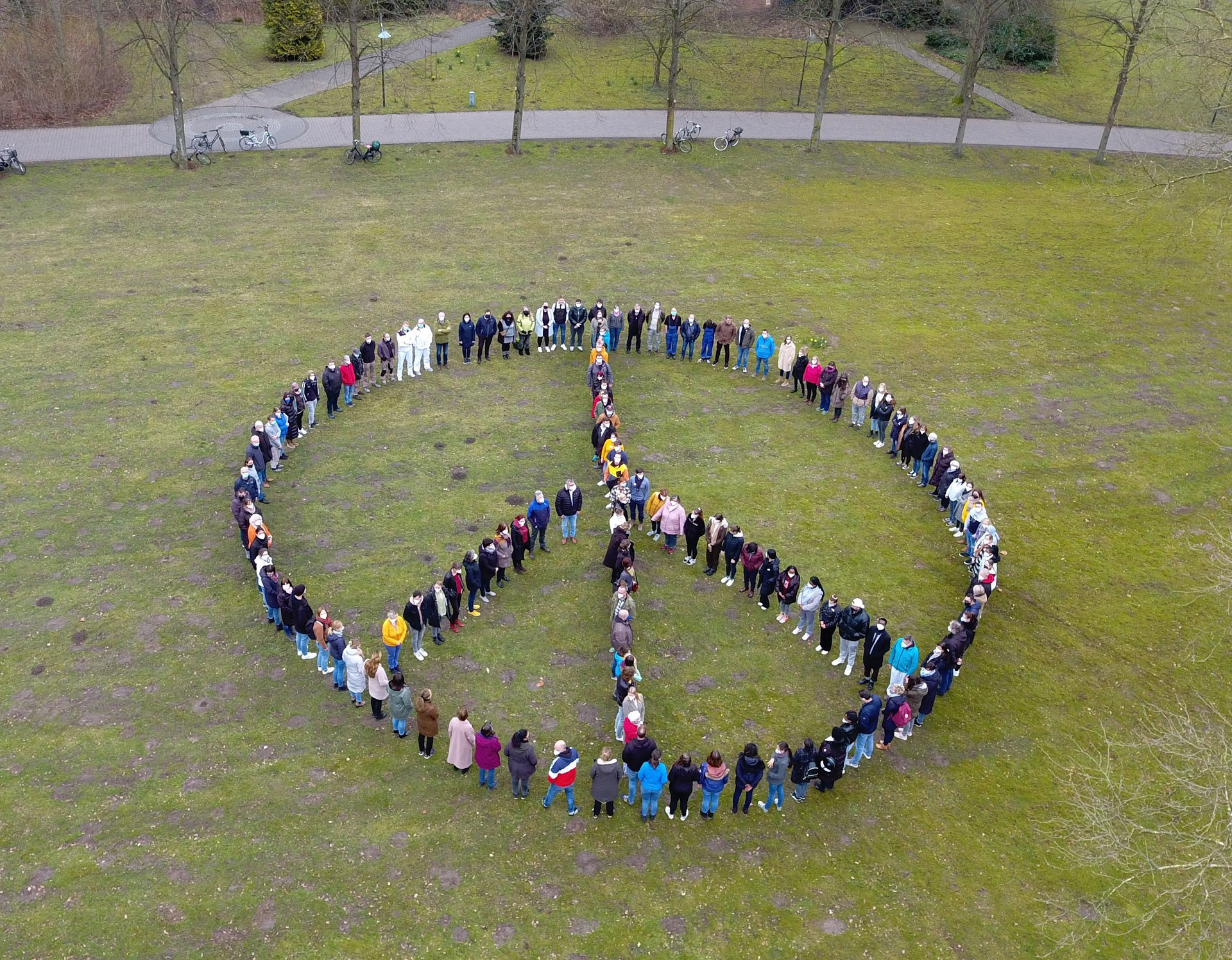 Titelfoto des Fortbildungsprogramms: Auf einer grünen Wiese stehen 150 Menschen, die das Friedenszeichen bilden.