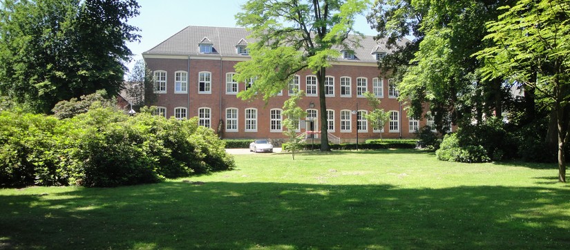Verwaltungsgebäude der Klinik zeigt ein rotes, älteres, zweistöckiges Gebäude im grünen Klinikpark, vor dem Gebäude parkt ein Auto