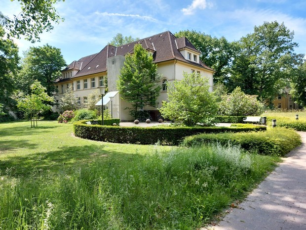 Foto zeigt ein zweistöckiges, weißes Gebäude inmitten eines grünen Parks im Sommer