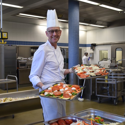 Ein Koch mit weißer Kleidung und weißer Kochmütze hält zwei silberfarbene Tabletts mit Pizza in der Hand