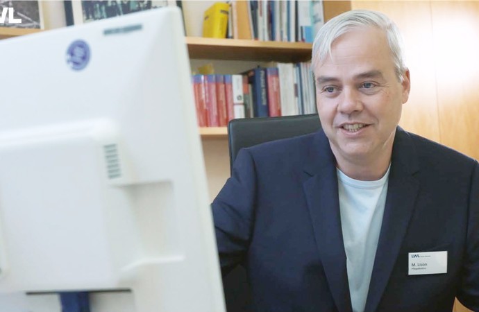 Ein Mann im dunkelblauen Jacket und weißem Shirt schaut in einen PC-Monitor, im Hintergrund Bücherregal