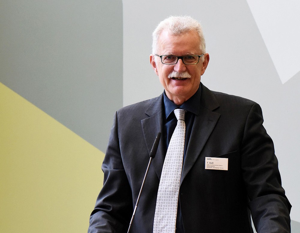 Ein Mann im Anzug steht vor einem grau-grünen Hintergrund