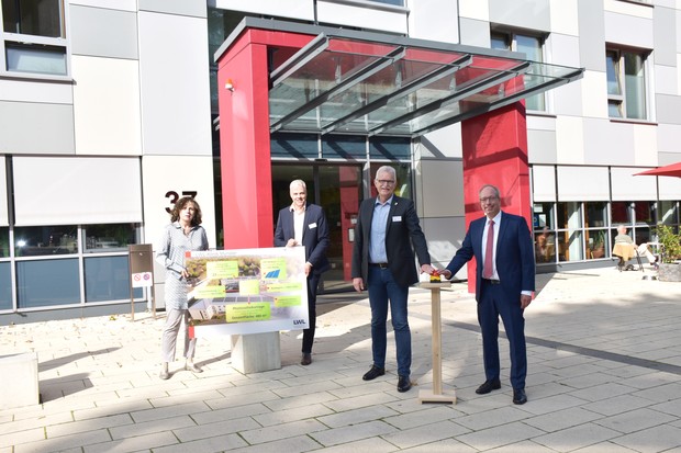 Eine Frau und drei Männer stehen vor dem roten Eingang des Klinikhauptgebäudes und halten ein Schild mit Daten zur neuen Photovoltaikanlage