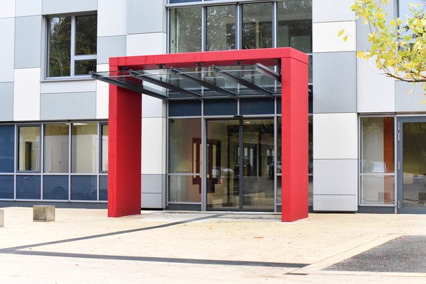 Roter, eckiger, moderner Torbogen umrahmt den Haupteingang des Klinikgebäudes