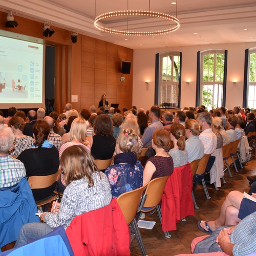 Viele Menschen sitzen in einem großen Saal, vor ihnen steht ein Mann mit einem Mikrophon.