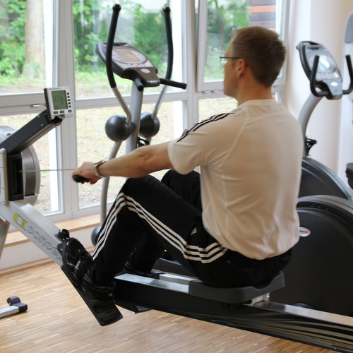 Ein Mann trainiert mit schwarzer Hose und einem weißen Shirt in einem Fitnessraum, sitzend auf einem Ergometer