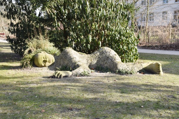 Eine Steinskulptur in Form eines auf der Seite liegenden Menschen liegt eingebettet in eine Rasenfläche unter einem Baum
