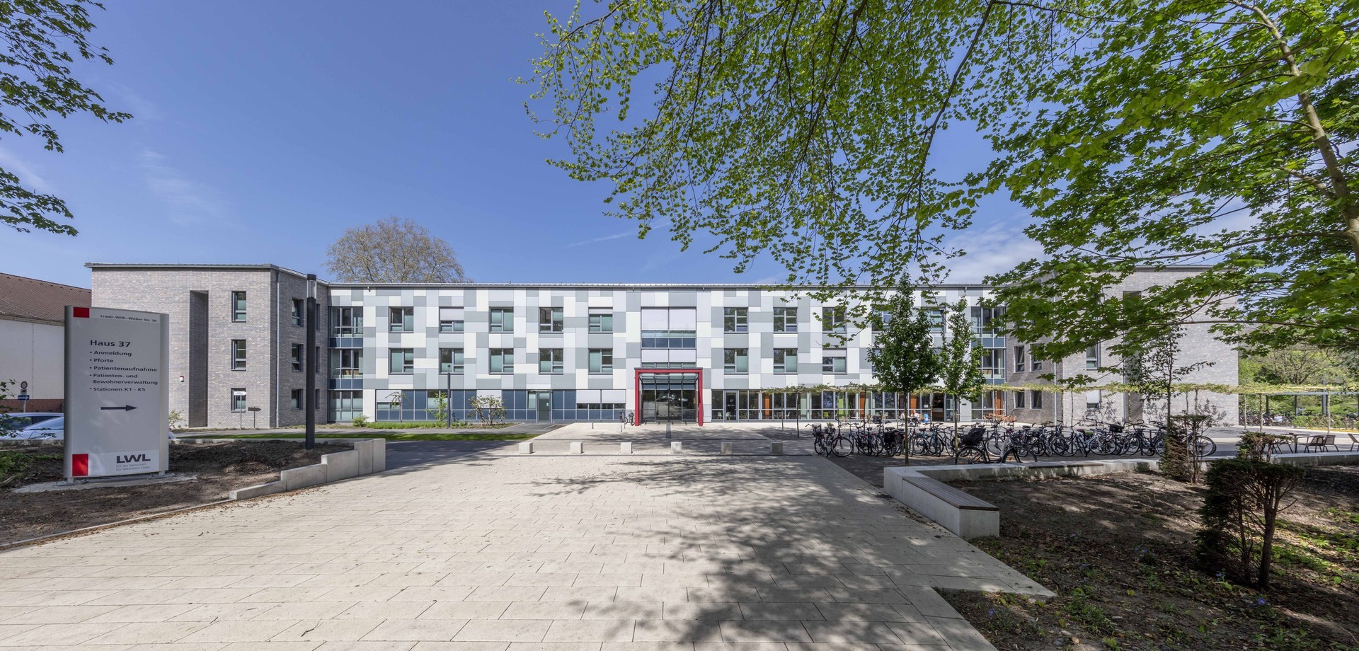 Hauptgebäude der LWL-Klinik Münster. Ein zweistöckiges modernes Gebäude aus hellem, grauen Stein, die große Eingangstür ist mit roten Steinen umrahmt