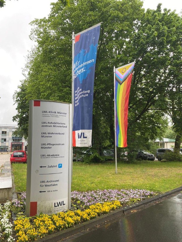 Foto zeigt zwei Fahnenstangen vor dem Hauptgebäude der LW-Klinik Münster, die eine Flagge zeigt die Pride-Flag, die andere die blaue LWL-Fahne
