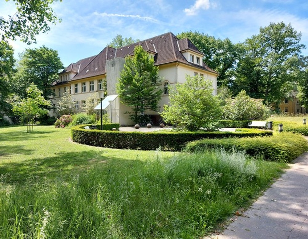 Im Vordergrund grüner Rasen, dahinter ein zweistöckiges Gebäude aus weißem Stein mit einem roten Dach, eingebettet in den grünen Park der Klinik