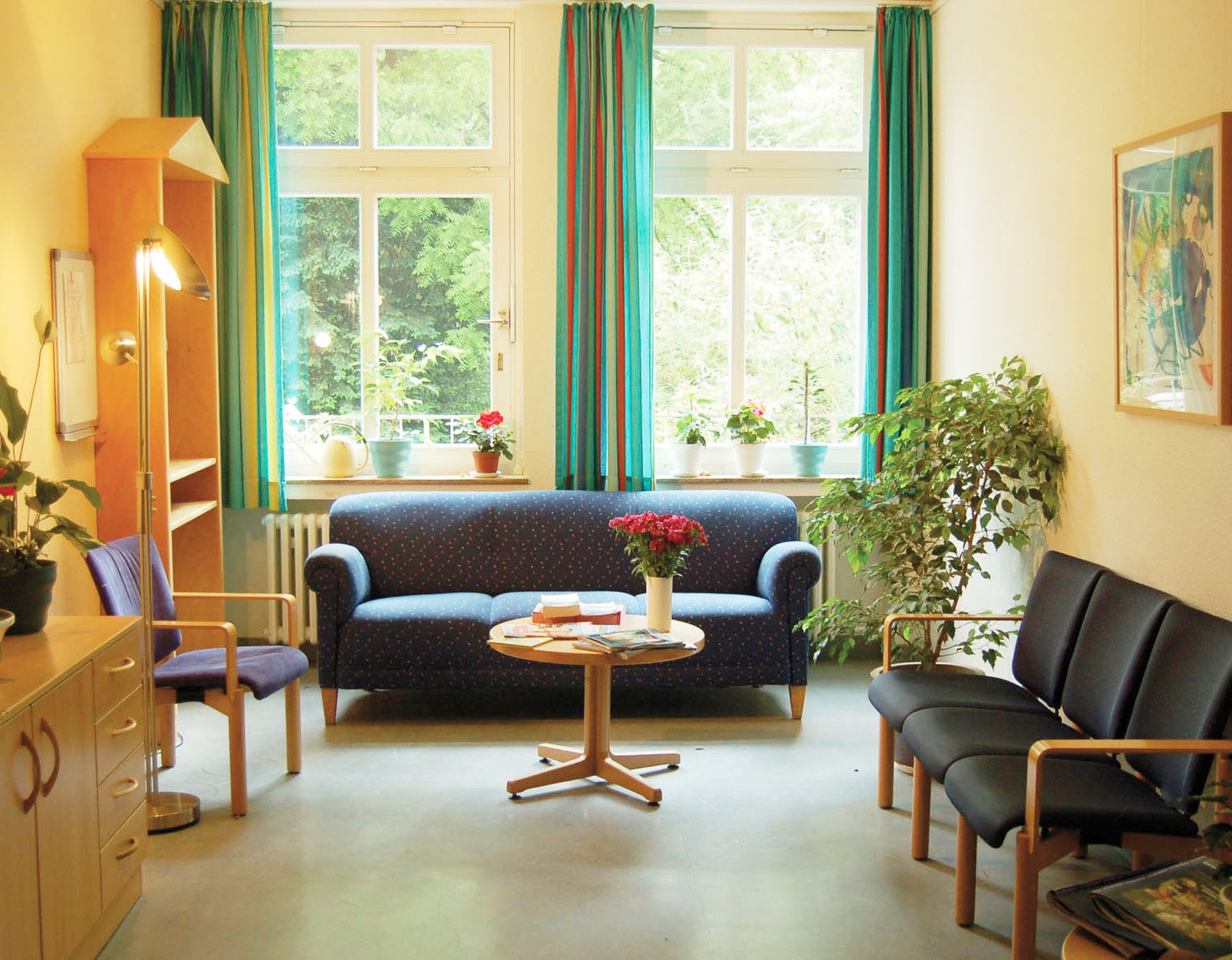 Aufenthaltsraum: Blaue Polstermöbelsitzgruppe vor einem Fenster mit grünen Vorhängen, auf dem Tisch ein Blumenstrauß und verschiedene Bücher