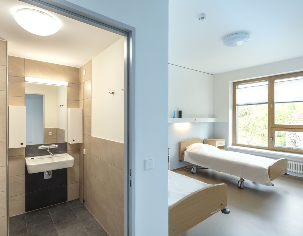 Patientenzimmer: Links ein Badezimmer mit Dusche, Waschbecken und Spiegel, die Wände sind hellbraun, rechts stehen zwei Betten vor hellblauen Wänden
