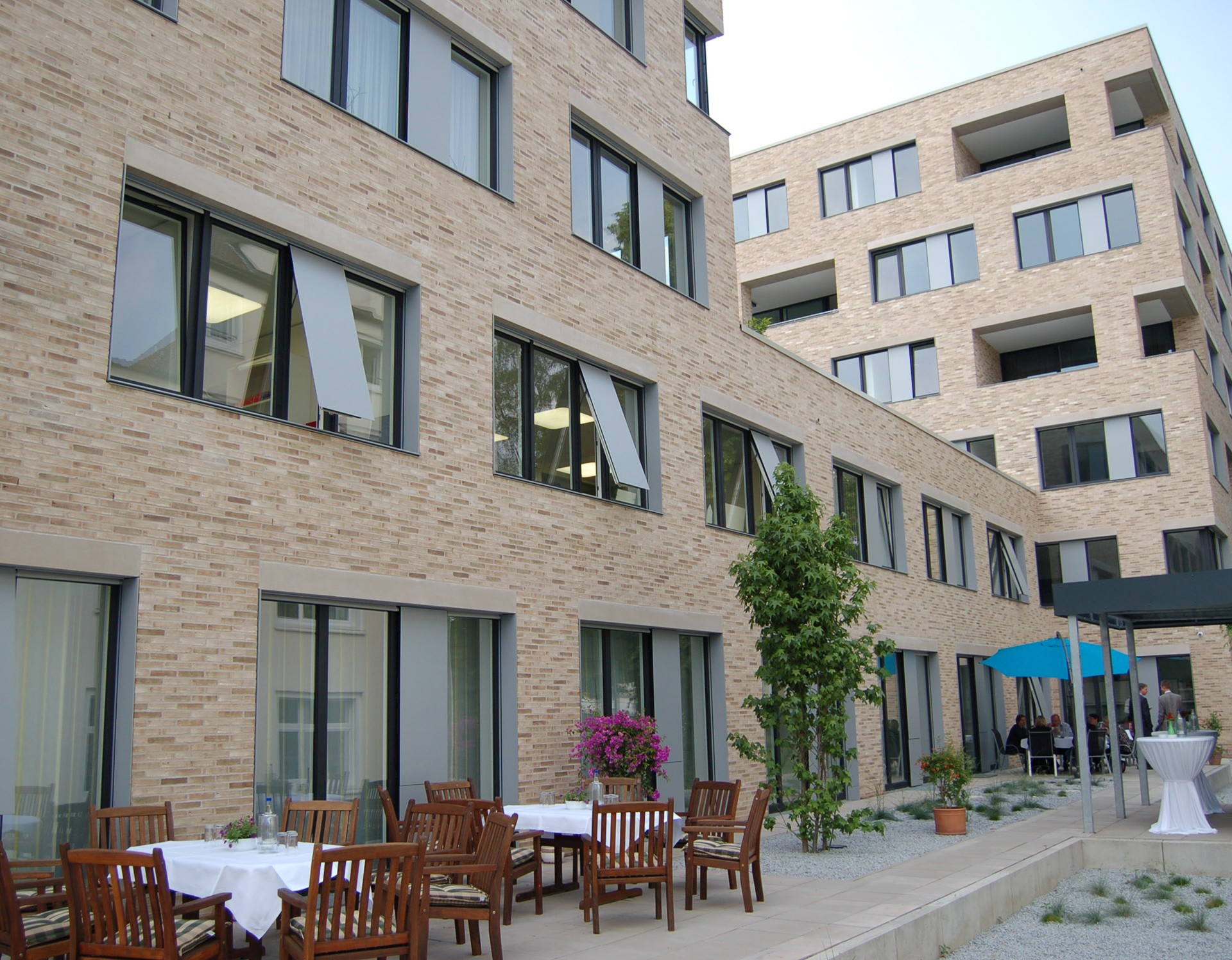Mehrstöckiges modernes, hell-beiges Gebäude, auf der Terrasse davor stehen einige braune Stühle und Tische mit einem blauen Sonnenschirm