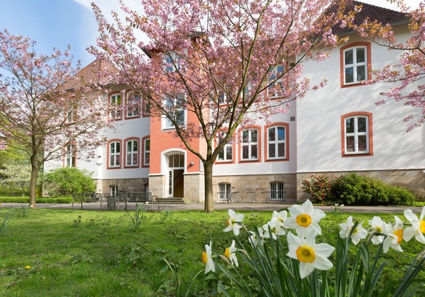 Zweistöckiges, rot-weißes Gebäude der Pflegeschule, im Vordergrund grüner Rasen und gelbe Narzissen Zweistöckiges, rot-weißes Gebäude der Pflegeschule, im Vordergrund grüner Rasen und gelbe Narzissen