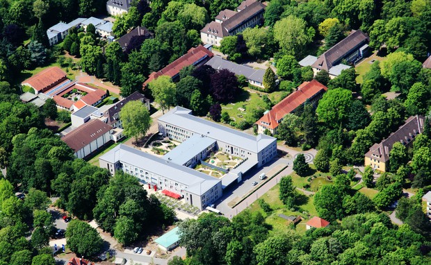 Luftbildaufnahme auf das grüne Klinikgelände der LWL-Klinik Münster, in der Mitte das große zweistöckige Hauptgebäude aus hellgrauem Stein