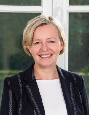 Eine Frau mit hellem Oberteil und dunklem Blazer lächelt.