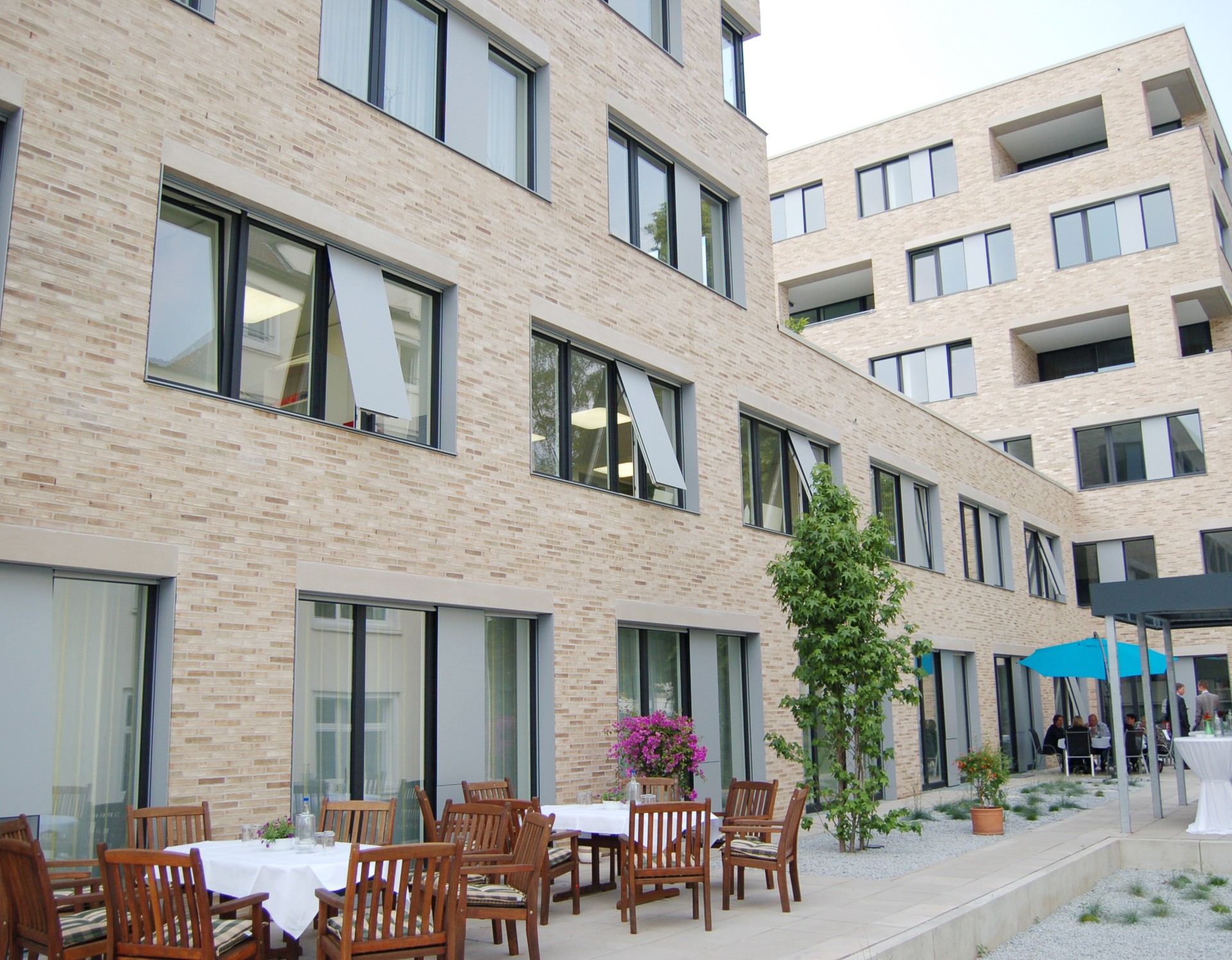 Das moderne hell-beige Gebäude an der Zumsandestraße, davor eine Terrasse mit zwei hölzernen Tischgruppen, daneben ein grüner Strauch