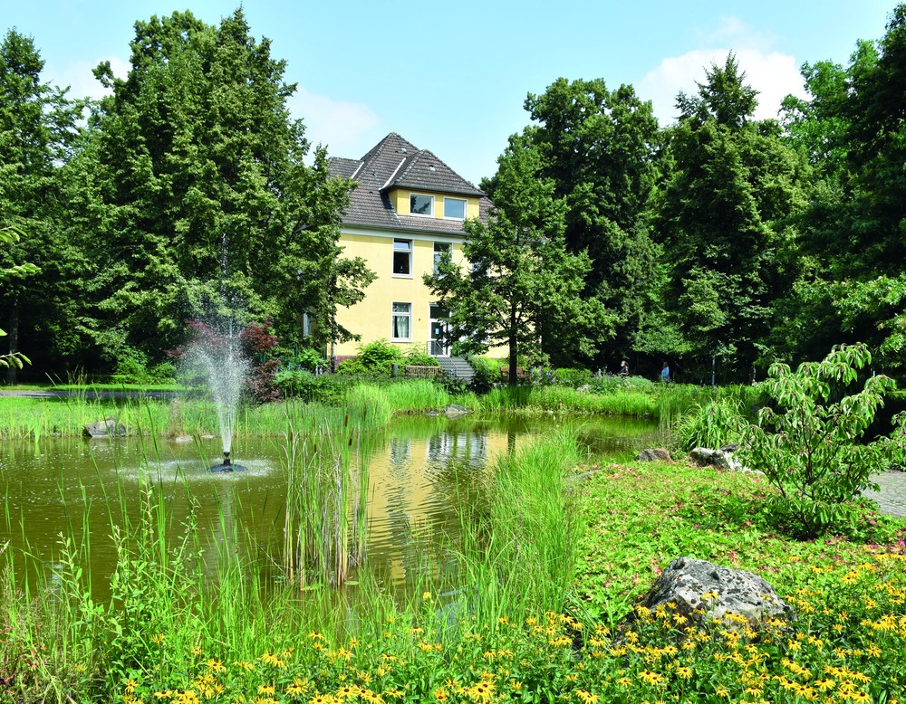 Foto zeit das Gebäude, Haus 24, ein hellgelbes, zweistöckiges Gebäude mit grauem Dach, im Vordergrund Teich mit grünen Sträuchern