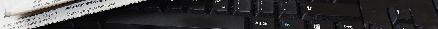 Auf einem hellen Holztisch liegt eine schwarze Tastatur, darauf vier Tageszeitungen