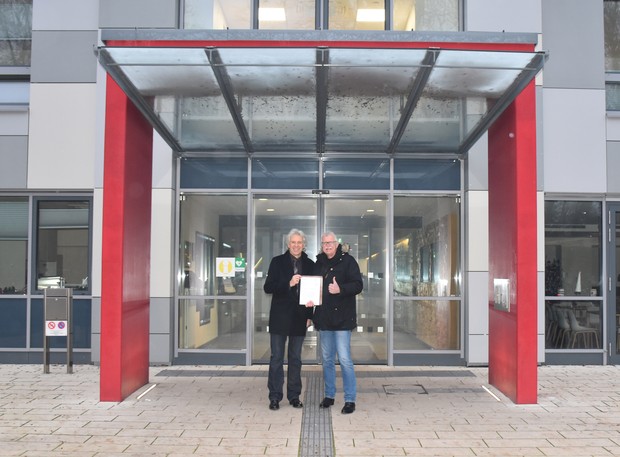 Zwei Männer stehen vor einer aus Glas bestehenden großen Eingangstür eines Gebäudes und halten ein Papier in der Hand.